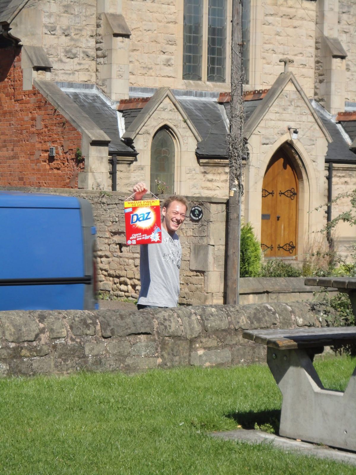 Daz, a legend, holding up a box of Daz washing powder in front of a church.
